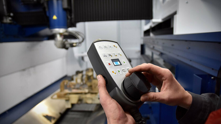 Laserschneiden - TruLaser Cell 8030 Der Fokus dieses Bildes liegt auf den Händen, die sorgfältig eine Bediensteuerung einer Laserschneidmaschine halten. Im Hintergrund, leicht verschwommen, um die Aufmerksamkeit auf die Hände und die Steuerung zu lenken, ist der Laserschneidkopf zu erkennen. Dieser ist gerade in Aktion oder bereit, in Betrieb genommen zu werden, was die Präzision und Technik hinter dem Laserschneiden unterstreicht. Die Darstellung fängt ein Moment der Konzentration und Fachkenntnis ein, der für die Bedienung solch spezialisierter Maschinerie erforderlich ist.