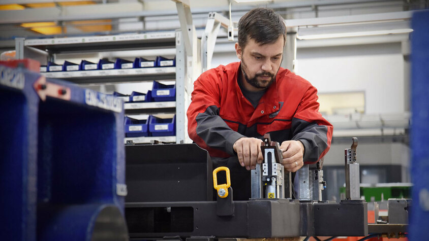Expertise in der Umformtechnik - Werkzeugherstellung, Vorrichtungsbau und mehr Das Bild zeigt einen Werkzeugbautechniker von weba, der mit Konzentration eine Prüfteillehre in einer produktionsnahen Umgebung zusammenbaut. Die Szene ist von Werkzeugen, Maschinen und anderen Arbeitsutensilien umgeben. Der Techniker arbeitet präzise und sorgfältig, um sicherzustellen, dass die Lehre korrekt montiert wird. Die Atmosphäre strahlt Professionalität und Effizienz aus, während der Techniker die Lehre für den Einsatz in der Produktion vorbereitet. Das Bild verdeutlicht webas übergreifende Expertise in der Umformtechnik.