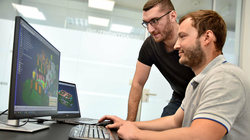 Jobs in Steyr - Berufseinstieg bei weba Werkzeugbau Zwei junge Konstrukteure bei weba Werkzeugbau vertieft in die Betrachtung einer 3D-Konstruktion eines Warmumformwerkzeugs. Einer der Konstrukteure sitzt vor einem PC, auf dessen Bildschirm die detaillierte 3D-Konstruktion lebhaft dargestellt ist, während der andere neben ihm steht und gemeinsam auf den Bildschirm blickt. Sie wirken konzentriert und engagiert, als würden sie wichtige Aspekte des Designs diskutieren. Diese Szene veranschaulicht Teamarbeit und den Einsatz modernster Technologien im Entwicklungsprozess bei weba, unterstreicht die Bedeutung von Kollaboration und Innovation in der Konstruktionsarbeit. Das Bild wird symbolisch für Jobs in Steyr für Berufseinsteiger verwendet.