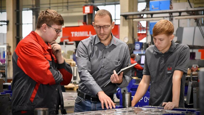 Stanz- und Umformwerkzeuge - Lehrlingsausbildung bei weba Werkzeugbau Das Bild zeigt den Lehrlingsausbilder, der zwei Lehrlingen in der Werkzeugbautechnik in einer Produktionsumgebung ein Stanz- und Umformwerkzeug erklärt. Er zeigt auf bestimmte Teile des Werkzeugs und erklärt die Funktionen und den Zweck jeder Komponente. Die Lehrlinge hören aufmerksam zu und stellen Fragen, während sie lernen. Die Szene vermittelt den Eindruck von Wissensaustausch und praktischer Schulung in einem professionellen Arbeitsumfeld.