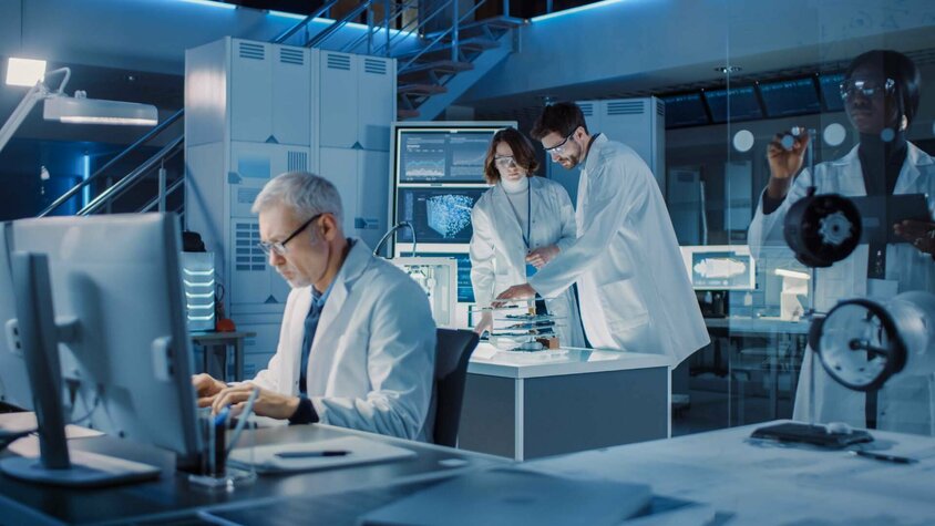 Innovationen in der Umformtechnik - Forschung und Entwicklung schafft Vorsprung Ein diverses Team von Industriewissenschaftlern und Ingenieuren in weißen Kitteln arbeitet konzentriert in einem Forschungslabor. Sie nutzen Computer und kommunizieren miteinander, während sie an der Entwicklung schwerer Maschinen arbeiten. Im Hintergrund sind technische Zeichnungen sichtbar, die die Komplexität ihrer Projekte verdeutlichen. Die Szene strahlt Professionalität und Teamarbeit aus, mit einem Fokus auf Innovation in der Umformtechnik und im Maschinenbau.