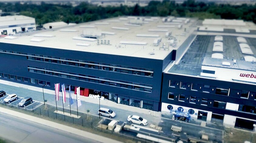 weba Olomouc - Serienproduzent und Experte in der Warmumformung Luftaufnahme des Produktionswerkes von weba für Warmumformung und Serienfertigung in Olomouc. Die Aufnahme zeigt die anthrazitfarbene Gebäudefront und dahinter eine riesige Produktionshalle. Vor dem Werk wehen die österreichische, tschechische, europäische sowie die Unternehmensflaggen.