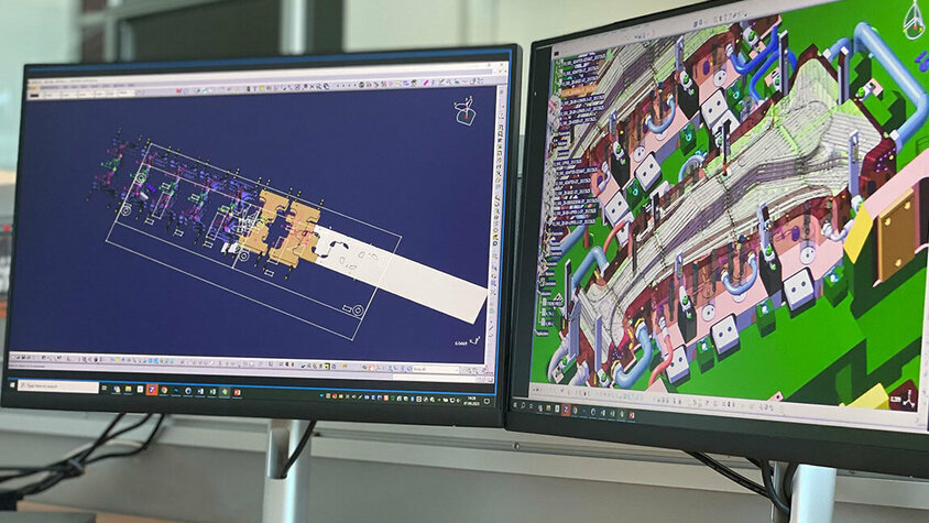 CAD Konstruktion - Innovatives Werkzeugdesign Zwei PC-Bildschirme, einer zeigt ein detailliertes Close-up einer CAD Konstruktion eines Warmumformwerkzeuges zur Produktion von B-Säulen, während der andere Bildschirm den Transferaufbau eines Werkzeuges darstellt.
