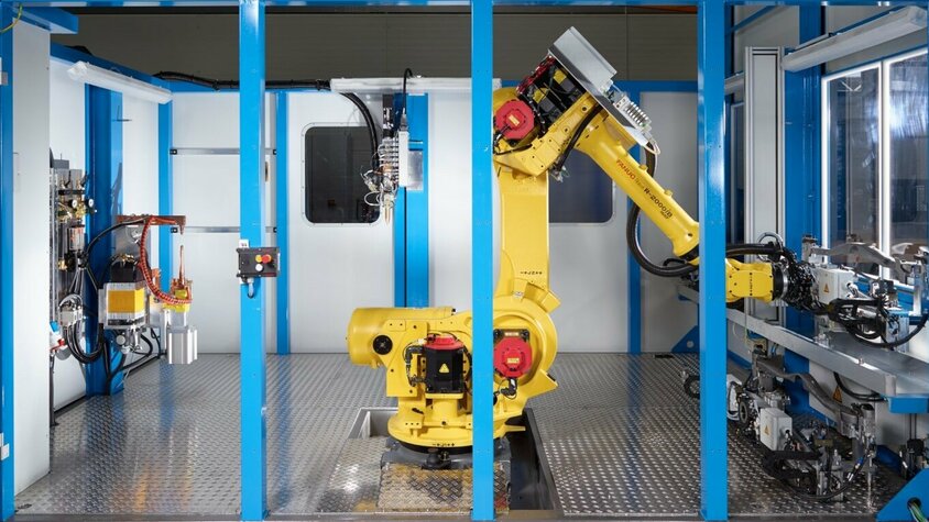 Fügetechnik Klebeanlage Eine moderne Fügetechnik-Anlage mit einem gelben FANUC-Roboterarm in einem abgeschirmten Arbeitsbereich. Der Roboter ist zentral in der Anlage positioniert und bereit für automatisierte Klebeaufgaben. Umgeben von blauen Rahmenstrukturen zeigt die Szene eine hochmoderne Fertigungsumgebung mit präzisen mechanischen Komponenten und fortschrittlichen Steuergeräten. Diese Anlage steht für Präzision, Effizienz und Automation in der Fügetechnik.