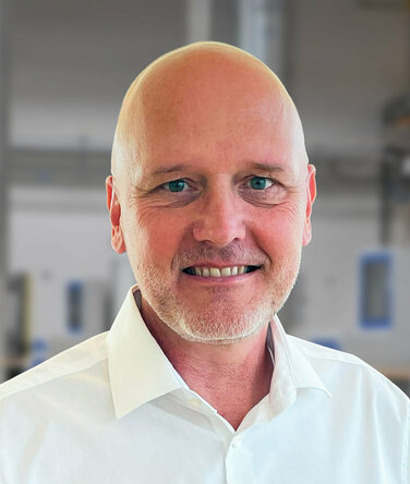 Andreas Mehlmayer - Vertriebsleitung Porträt von Andreas Mehlmayer in einem modernen Industrieumfeld. Er trägt ein weißes Hemd und lächelt in die Kamera. Im unscharfen Hintergrund sind helle Fertigungseinrichtungen und Maschinen erkennbar – typisch für eine präzise und saubere Produktionsumgebung.