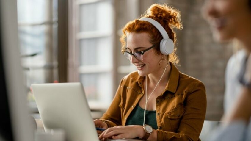 Praktikum Jobs in Steyr - Karrierestart bei weba Lächelnde kreative junge Frau hört Musik mit Kopfhörern, während sie im Büro an einem Computer bei ihrem Praktikum Job in Steyr bei weba arbeitet.