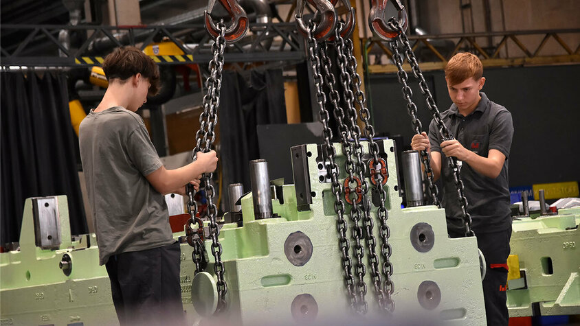 Stanz- und Umformwerkzeuge - Lehrlingsausbildung in der Metalltechnik bei weba Werkzeugbau Das Bild zeigt zwei Lehrlinge in der Werkzeugbautechnik, wie sie ein Umformwerkzeug mit einem Kran spannen. Beide sind konzentriert bei der Arbeit und zeigen Teamarbeit beim sicheren Anheben des Werkzeugs. Die Szene vermittelt den Eindruck von Lernbereitschaft und praktischer Erfahrung im Umgang mit schweren Werkzeugen.