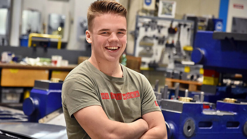 Jobs in Steyr - Lehre bei weba Werkzeugbau als Werkzeugbautechniker Ein Lehrling bei weba Werkzeugbau, fotografiert in der Produktionshalle, lächelt direkt in die Kamera. Er trägt ein T-Shirt mit der Aufschrift 'We are WEBA', was seinen Stolz und seine Zugehörigkeit zum Unternehmen zum Ausdruck bringt. Im Hintergrund sind mehrere Werkbänke und Teile eines dunkelblauen Umformwerkzeugs sichtbar, was auf die praktische und lernintensive Umgebung hinweist, in der der Lehrling ausgebildet wird. Das Bild strahlt Jugendlichkeit, Optimismus und das Engagement des Lehrlings für seine Ausbildung aus, während es gleichzeitig die unterstützende und lehrreiche Atmosphäre bei weba hervorhebt. Das Bild steht symbolisch für Lehrstellen Jobs in Steyr bei weba Werkzeugbau.