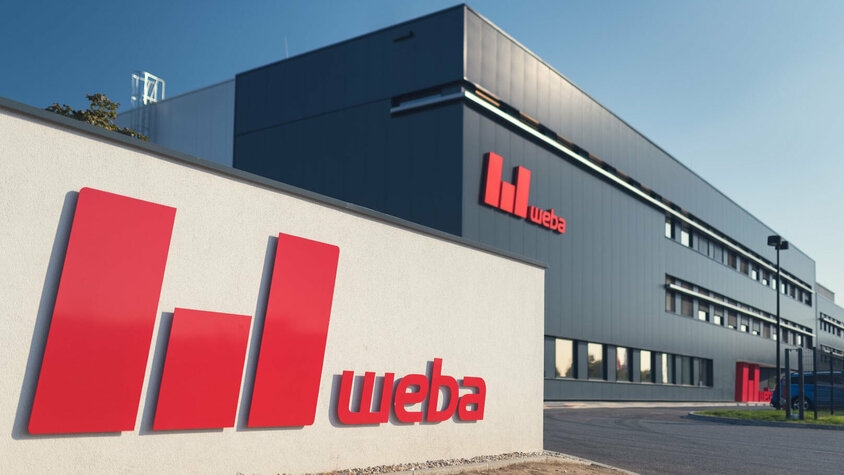 Expertise in der Umformtechnik - Produktionsstandort weba Olomouc Das Bild zeigt die Außenansicht des Produktionsstandorts weba Olomouc. Die Gebäudefront ist anthrazitgrau gestaltet, was einen modernen und professionellen Eindruck vermittelt. Vor der Einfahrt ist das rote weba-Logo auf der Mauer deutlich sichtbar. Die Szene strahlt Stabilität und Markenidentität aus, während das Logo Unternehmen und Standort identifiziert.