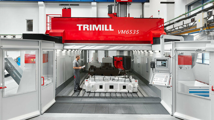 Presshärten - Hohe Flexibilität in der Herstellung durch umfangreichen Maschinenpark bei weba Werkzeugbau in Olomouc Das Bild zeigt eine große Zerspanungsmaschine der Marke Trimill mit geöffneter Tür, die einen Blick auf den Frästisch ermöglicht. Auf dem Frästisch ist ein großer Guss zu sehen, der auf die Bearbeitung wartet. Die Szene vermittelt den Eindruck von industrieller Fertigung und Präzisionsbearbeitung in einer Werkstattumgebung.