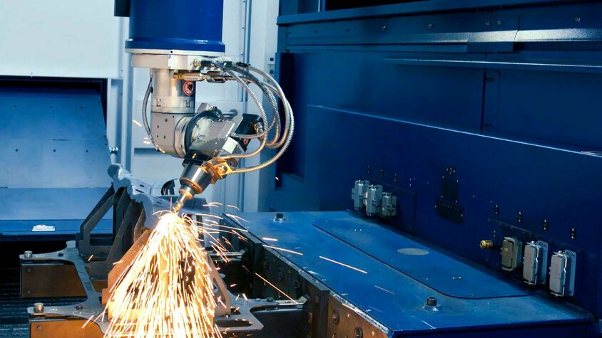 Hochpräzises 3D-Laserschneiden von Metall Ein Laserschneidkopf schneidet mit hoher Präzision ein Metallwerkstück in einer industriellen Maschine. Funken fliegen in verschiedene Richtungen, während der Laser das Material bearbeitet. Die Umgebung ist in einem industriellen Blau gehalten und die Maschine ist mit mehreren technischen Komponenten ausgestattet.
