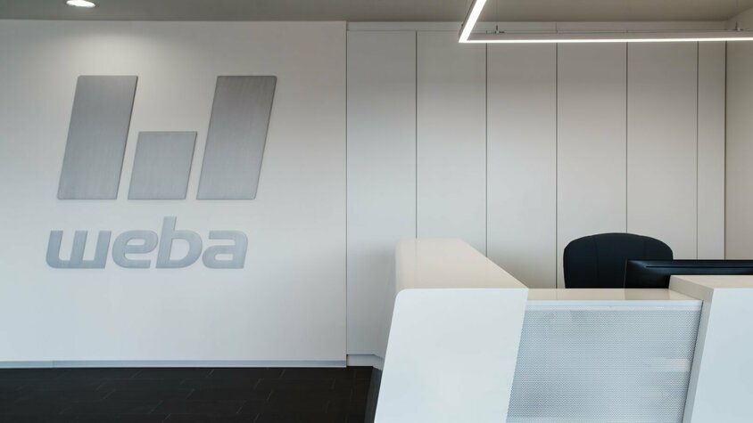 weba Olomouc - High-Tech Werkzeugbau im modernen Design Moderne weiße Empfangstheke im weba-Standort Olomouc, mit dem weba-Logo in gebürstetem Aluminium an der Wand dahinter.