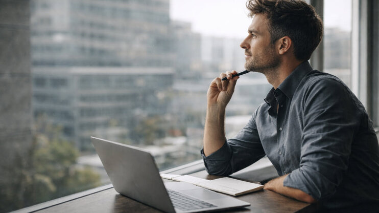 A working professional sits at a table with a laptop and notebook, thoughtfully looking out of the window of a modern office building.