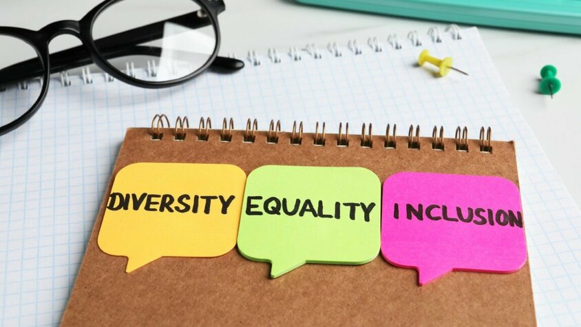 Arbeitskultur und Karriere in Steyr - Inklusivität, Diversität und Gleichheit Hinterseite eines Blocks mit drei bunten Post-its, auf denen die Wörter 'Diversity, Equality und Inclusion' stehen. Daneben liegen eine schwarze Rahmenbrille, eine türkisfarbene Tastatur sowie zwei Pinnadeln in grün und gelb.