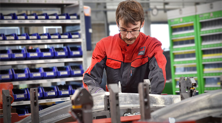 Das Bild zeigt einen Werkzeugbautechniker, der konzentriert ein Presshärten-Werkzeug für die Umformung von Strukturbauteilen zusammenbaut. Im Hintergrund sind Regale mit Hilfs- und Betriebsstoffen zu erkennen, die unscharf sind und den Arbeitsbereich des Technikers ergänzen. Die Szene vermittelt den Eindruck von Präzision und Organisation in der Fertigungsumgebung.