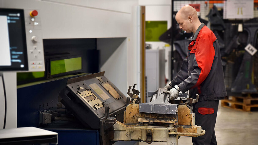 Präzisen 3D-Laserschneiden eines Metallwerkstücks Ein Mitarbeiter bedient eine Laserschneidmaschine in einer Produktionshalle. Er hält ein Werkstück aus Metall, das gerade in die Maschine zum Laserschneiden eingesetzt wird. Die Arbeitsumgebung ist sauber und gut organisiert, mit verschiedenen Werkzeugen und Materialien im Hintergrund.