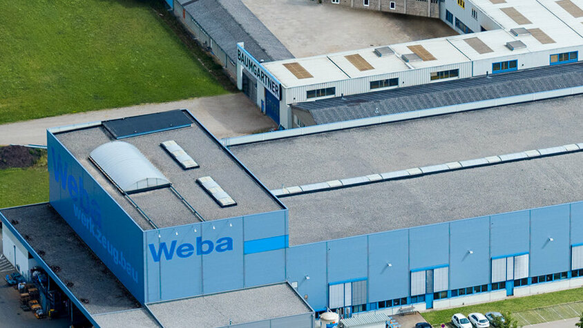Stanz- und Umformwerkzeuge - Expertise von weba Werkzeugbau in Dietach Die Luftaufnahme zeigt das beeindruckende Headquarters und Entwicklungszentrum von weba in Dietach. Die Produktionsstätte erstreckt sich über das Gelände, während im Vordergrund die Pressenhalle mit der deutlich sichtbaren Beschriftung "WEBA" zu sehen ist. Die Architektur wirkt modern und funktional, während die Umgebung von Grünflächen und Straßen umgeben ist. Die Szene vermittelt einen Eindruck von Größe und Professionalität, während das "WEBA"-Logo die Identität des Unternehmens betont.