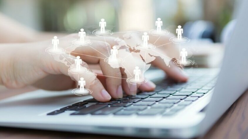 weba Werkzeugbau - Social Media Hände arbeiten an einem Laptop, darüber ist die Weltkarte in hoher Transparenz gelegt, mit Strichmännchen, die sich über die Kontinente vernetzen.