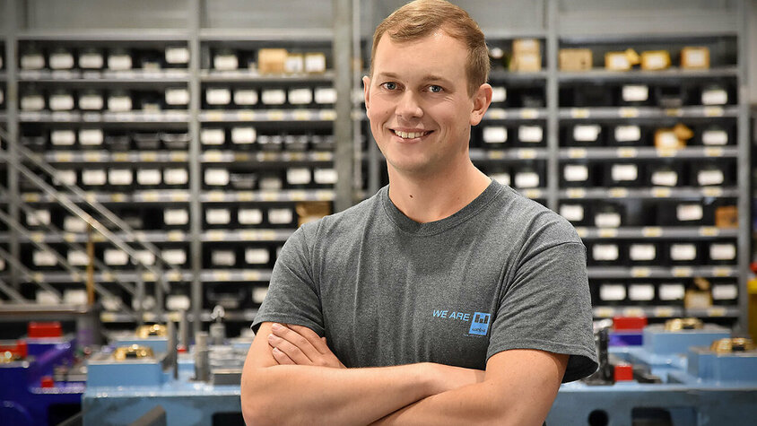 Jobs in Steyr - Praktikum bei weba Werkzeugbau Ein Werkzeugbautechniker von weba Werkzeugbau, fotografiert in der Produktionsumgebung, blickt direkt und mit einem stolzen Lächeln in die Kamera. Er steht in einer selbstbewussten Pose, die seinen Stolz auf seine Arbeit unterstreicht. Im Hintergrund sind ordentlich angeordnete Betriebsmittelregale und ein markantes blaues Umformwerkzeug zu sehen, was auf die gut organisierte und technologisch fortschrittliche Arbeitsumgebung hinweist. Das Bild vermittelt ein Gefühl von Professionalität und Zufriedenheit im Arbeitsumfeld, und hebt die persönliche Verbindung des Technikers zu seiner Arbeit und seinem Arbeitsplatz hervor. Das Bild steht symbolisch für Praktikum-Jobs in Steyr.