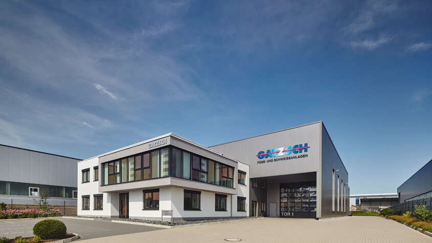 Gatzsch Schweißtechnik - Experte für High-Tech Fügeanlagen Die Außenansicht des Werkes Gatzsch Schweißtechnik GmbH zeigt ein strahlendblaues Himmel und das Verwaltungsgebäude im Fokus. Dahinter erstreckt sich die Produktionsanlage, auf der ein großes Logo mit den Worten 'Gatzsch Füge- und Schweissanlagen' deutlich sichtbar ist.