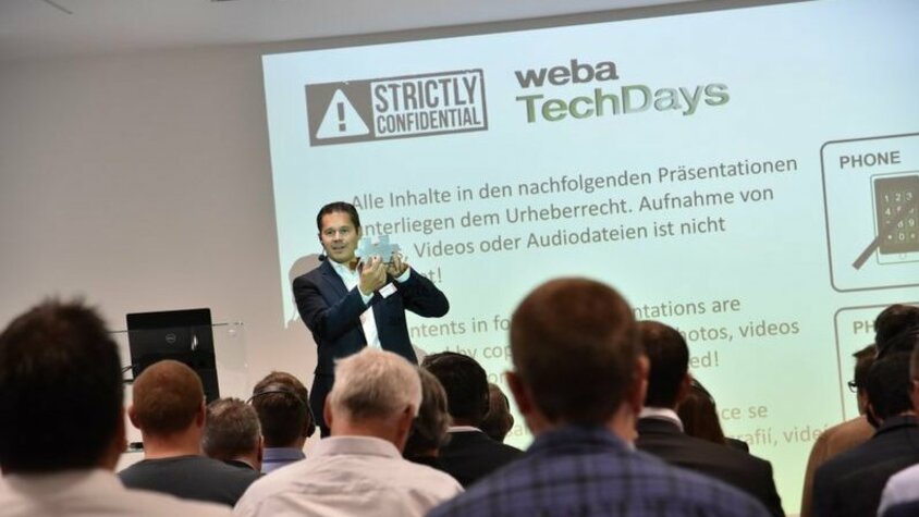weba Werkzeugbau TechDays - Die Welt des Werkzeugbaus Geschäftsführer Hannes Feuerhuber von weba Werkzeugbau begrüßt das Publikum der Werkzeugbaukonferenz auf der Bühne, während im Hintergrund die Geheimhaltungsrichtlinien projiziert werden.