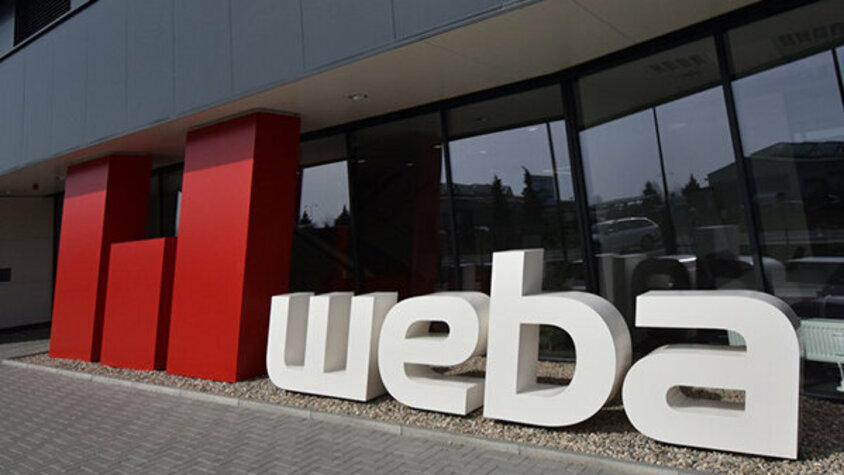 weba Olomouc - Experte in der Warmumformung und Werkzeugbau Fotoaufnahme vor dem Eingang des Experten im Werkzeugbau und der Warmumformung weba Olomouc: Anthrazitfarbene Gebäudefront mit Fensterfront, davor das weba-Logo in Rot und Weiß in großen 3D-Buchstaben.