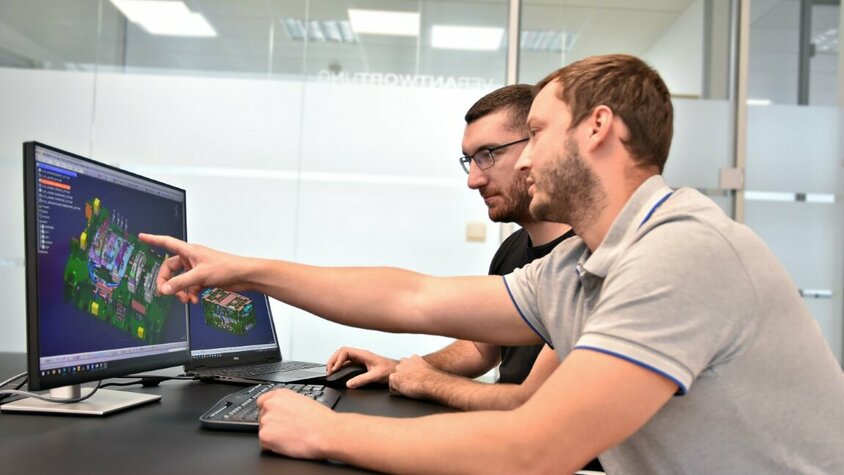Werkzeugherstellung - Werkzeugkonstruktion bei weba Werkzeugbau Zwei Personen sitzen vor zwei Computerbildschirmen, auf denen eine CATIA-Datei mit einer Werkzeugkonstruktion geöffnet ist. Eine der Personen zeigt auf ein spezifisches Detail des Werkzeugs auf dem Bildschirm, während sie offenbar die Konstruktion besprechen.