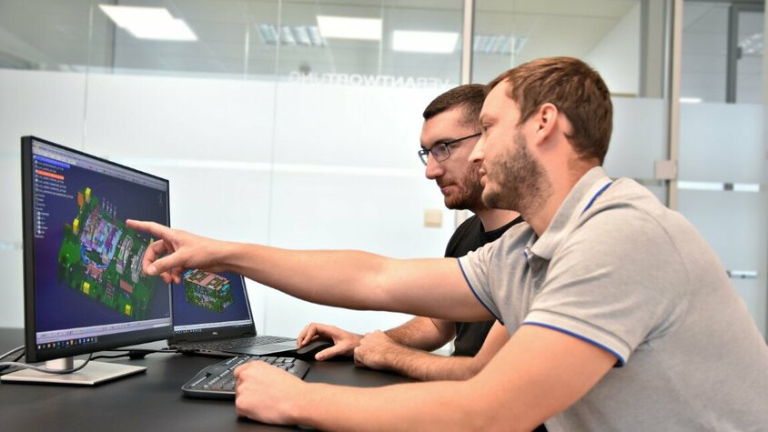 Werkzeugherstellung - Werkzeugkonstruktion bei weba Werkzeugbau Zwei Personen sitzen vor zwei Computerbildschirmen, auf denen eine CATIA-Datei mit einer Werkzeugkonstruktion geöffnet ist. Eine der Personen zeigt auf ein spezifisches Detail des Werkzeugs auf dem Bildschirm, während sie offenbar die Konstruktion besprechen.