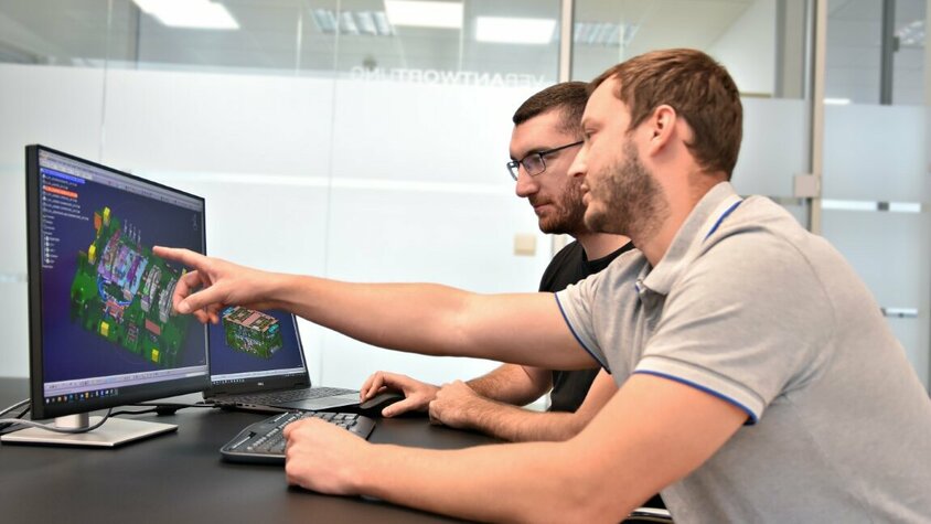 Werkzeugherstellung - Werkzeugkonstruktion bei weba Werkzeugbau Zwei Personen sitzen vor zwei Computerbildschirmen, auf denen eine CATIA-Datei mit einer Werkzeugkonstruktion geöffnet ist. Eine der Personen zeigt auf ein spezifisches Detail des Werkzeugs auf dem Bildschirm, während sie offenbar die Konstruktion besprechen.