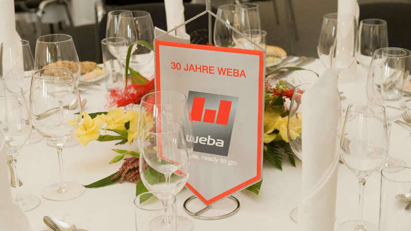 weba Werkzeugbau - Festliche Jubiläumsfeier Festliche Tischdeko beim VIP-Abend des 30-Jährigen Jubiläumsfests, mit Wimpel, auf dem das weba-Logo zu sehen ist, umgeben von elegantem Dekor.