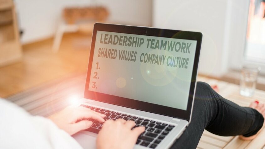 Arbeitskultur und Karriere in Steyr - Werte und Vision der weba Werkzeugbau Eine Frau sitzt auf dem Boden mit einem Laptop auf ihrem Schoß. Auf dem Bildschirm sind die Wörter 'Leadership, Teamwork, Shared Values und Company Culture' zu lesen, während das Licht auf dem Laptop reflektiert wird. Das Bild symbolisiert wertegebundene Arbeitskultur bei weba für deine Karriere in Steyr.