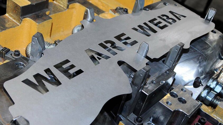 Jobs in Steyr - Karriereweg für Berfuserfahrene und Profis Eine Formplatine eines Stanzteils liegt in einem Umformwerkzeug. In die Platine ist der Schriftzug 'WE ARE WEBA' sorgfältig eingelasert, was auf die präzise Verarbeitung und die Identifikation mit dem Unternehmen hinweist. Das Bild wird Symbol für Teamwork und Begeisterung auf der Seite Jobs in Steyr für Berufserfahrene und Fachkräfte verwendet.