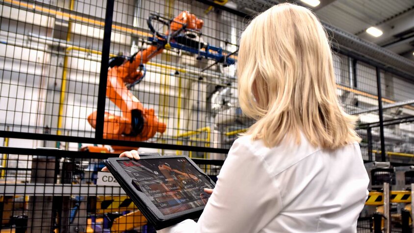 Žena stojí před velkým robotickým chapadlem na lince pro tváření za tepla v průmyslovém prostředí společnosti weba Werkzeugbau. V rukou drží tablet, na kterém sleduje data nebo zadává řídicí příkazy pro robota. Důraz je kladen na interakci mezi moderní technologií a lidským zásahem.