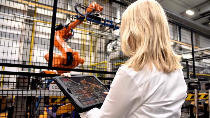 weba Werkzeugbau Digitale Qualitätskontrolle Eine Frau steht in einer industriellen Umgebung bei weba Werkzeugbau vor einem großen Robotergreifer einer Warmumformlinie. In ihren Händen hält sie ein Tablet, auf dem sie Daten überwacht oder Steuerbefehle für den Roboter eingibt. Der Fokus liegt auf der Interaktion zwischen moderner Technologie und menschlichem Eingreifen.