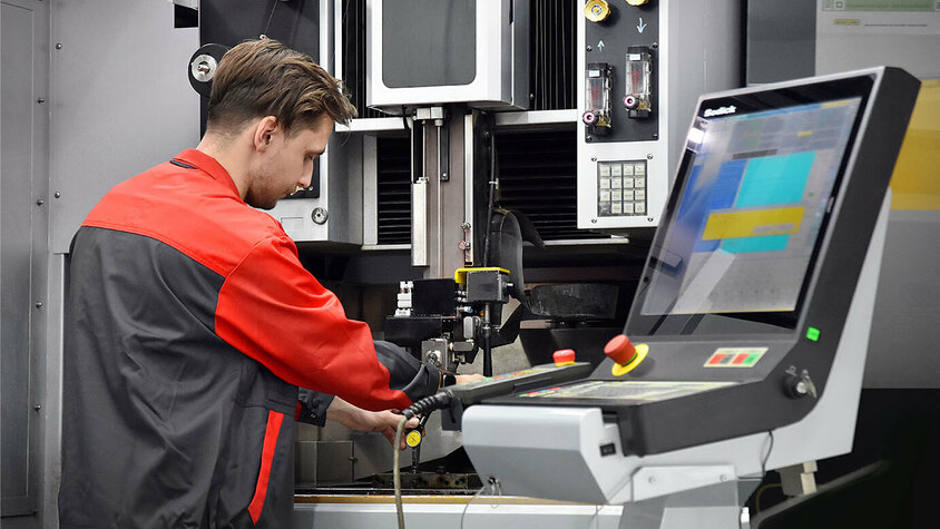 Erodieren im Presshärten - Expertise bei weba Olomouc Das Bild zeigt einen Werkzeugbautechniker von weba Olomouc, der konzentriert eine moderne Erodiermaschine bedient. Seine Hände sind geschickt damit beschäftigt, die Maschine zu steuern.