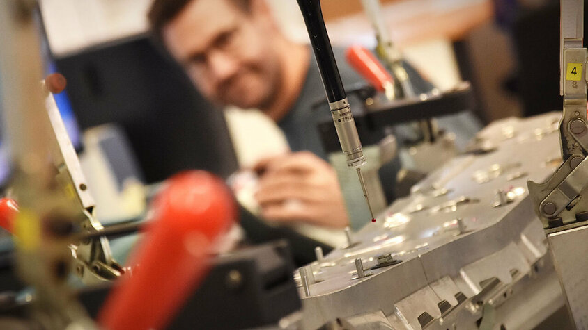 Messtechnik - 3D-Koordinantenmessung und taktile Messsysteme Nahaufnahme einer Koordinatenmessmaschine mit Fokus auf den Messtaster. Im Hintergrund ist der Messtechniker unscharf und verschwommen zu sehen.