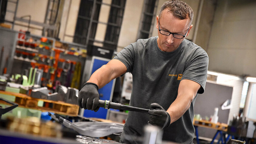 Stanz- und Umformwerkzeuge - Montage eines Stanzwerkzeuges bei weba Werkzeugbau Das Bild zeigt einen Werkzeugbautechniker, der konzentriert an der Montage eines Stanzwerkzeugs arbeitet. Seine Hände sind geschickt damit beschäftigt, die verschiedenen Komponenten des Werkzeugs zusammenzufügen. Im Hintergrund sind Werkzeugteile und Montagewerkzeuge zu erkennen. Die Szene vermittelt ein Gefühl von Handwerkskunst und Präzision bei der Herstellung hochwertiger Werkzeuge.