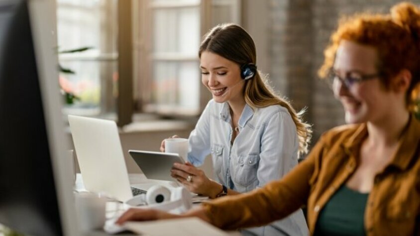 Berufseinstieg in Steyr - Stellenangebote Lächelnde weibliche Person mit Headset, die Kaffee trinkt und im Büro das Touchpad nutzt. Das Bild symbolisiert glücklichen Berufseinstieg in Steyr bei weba.