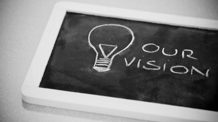 Arbeitskultur - Karriere in Steyr beim Technologieführer in der Warmumformung Eine auf dem Tisch liegende Tafel mit weißem Rahmen, auf der eine Glühbirne und der Text 'Our Vision' mit Kreide geschrieben sind. Das Foto symbolisiert innovative Arbeitskultur und herausragende Karriere in Steyr.