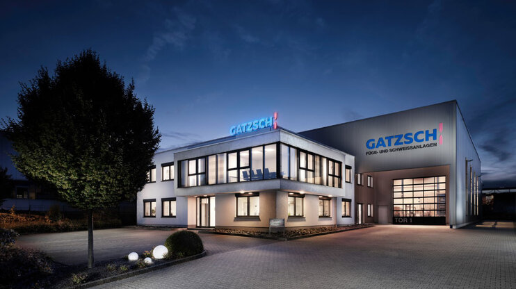 Exterior view of the Gatzsch company building at night, illuminated by modern outdoor lighting. The facility consists of an office building and a large production hall, prominently displaying the Gatzsch logo with the lettering “Joining and Welding Systems.” The windows emit a warm light that highlights the entrance area and surrounding premises. In the foreground, a well-maintained driveway with decorative landscaping is visible. The dark sky and atmospheric lighting create a professional and welcoming impression.