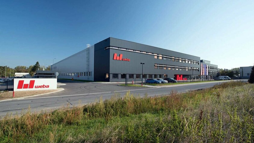 Produktionsstandort Olomouc - Experten für Presshärten und Serienfertigung Die Außenansicht des Produktionsstandorts weba Olomouc zeigt eine anthrazitgraue Gebäudefront, die einen modernen und professionellen Eindruck vermittelt. Das rote weba-Logo auf der Mauer vor der Einfahrt ist deutlich sichtbar. Die Szene strahlt Stabilität und Markenidentität aus, während das Logo Unternehmen und Standort identifiziert.