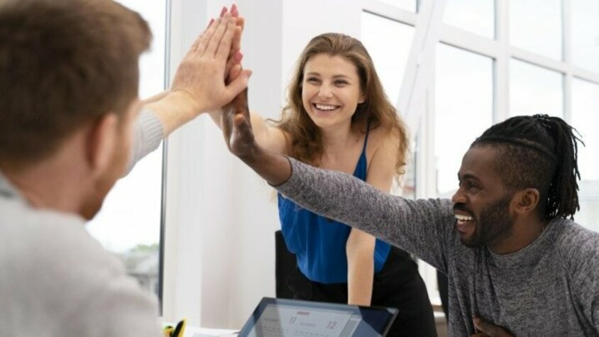 Jobs in Steyr - Deine Benefits bei weba Werkzeugbau Drei Personen sitzen lächelnd an einem Besprechungstisch und geben sich High-Five.