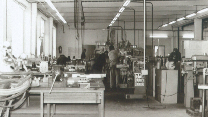weba Werkzeugbau - Die Erfolgsgeschichte Schwarz-weiße Archivaufnahme einer Werkstatt der weba aus den 80er Jahren, mit historischen Maschinen und Arbeitern in typischer Arbeitskleidung jener Zeit.