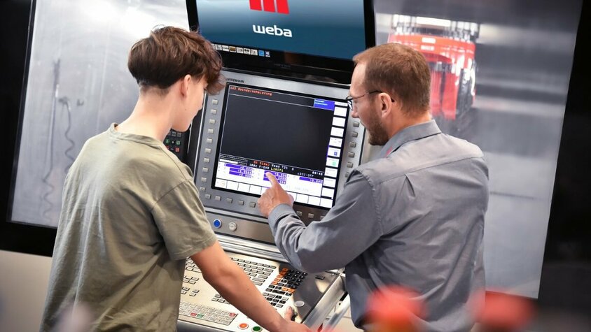 Lehre in Steyr - Ausbildung als Zerspanungstechniker Ein Ausbilder erklärt einem Lehrling in der Zerspanungstechnik die Bedienfunktionen an der Steuerung eines Bearbeitungszentrums. In der Lehre in Steyr bei weba Werkzeugbau liegt der Fokus auf praxisnaher Ausbildung direkt an Maschinen.