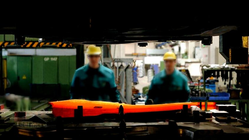 weba Werkzeugbau - Experte in der Warmumformung seit 1994 Ein durch den Spalt des Ober- und Unterteils einer Umformpresse aufgenommenes Bild fokussiert eine glühende Formplatine, die in ein Warmumformwerkzeug eingelegt ist. Im Hintergrund sind zwei Werkzeugbautechniker und die verschwommene Produktion zu sehen.