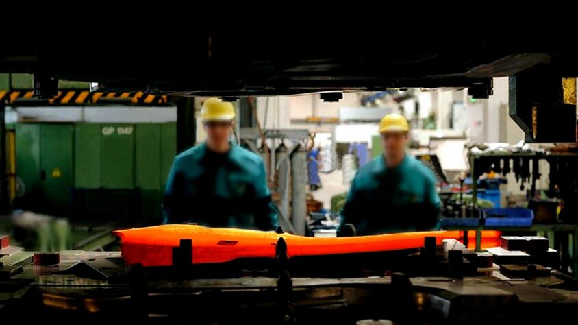weba Werkzeugbau - Experte in der Warmumformung seit 1994 Ein durch den Spalt des Ober- und Unterteils einer Umformpresse aufgenommenes Bild fokussiert eine glühende Formplatine, die in ein Warmumformwerkzeug eingelegt ist. Im Hintergrund sind zwei Werkzeugbautechniker und die verschwommene Produktion zu sehen.