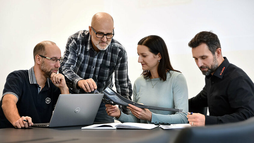 Jobs in Steyr - Karrieremöglichkeiten für Berufserfahrene bei weba Vier Personen sitzen an einem Besprechungstisch: Eine Person arbeitet an einem Laptop, eine andere betrachtet aufmerksam Unterlagen, während zwei weitere Personen sich angeregt über ein Stanzteil unterhalten. Die Szene zeigt eine konzentrierte und engagierte Arbeitsatmosphäre. Berufserfahrene und Profis finden bei weba ihren Traumjob in Steyr.