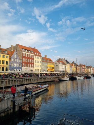 Nyhavn v Kodani s barevnými domy, loděmi na vodě a spoustou lidí podél promenády pod modrou oblohou.