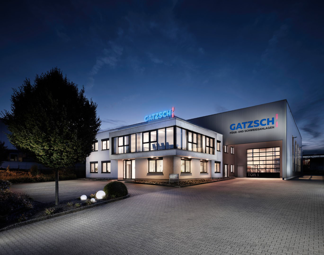 Exterior view of the Gatzsch company building at night, illuminated by modern outdoor lighting. The facility consists of an office building and a large production hall, prominently displaying the Gatzsch logo with the lettering “Joining and Welding Systems.” The windows emit a warm light that highlights the entrance area and surrounding premises. In the foreground, a well-maintained driveway with decorative landscaping is visible. The dark sky and atmospheric lighting create a professional and welcoming impression.