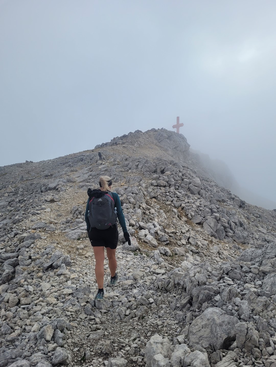 Bettina Gmainer s batohem stoupá po kamenité horské stezce, když se blíží k vrcholovému kříži. Obloha je zamlžená a atmosféra je syrová a dobrodružná.